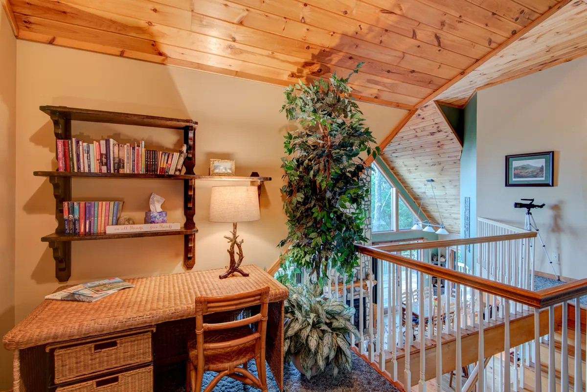 The image showcases a peaceful upstairs library nook. Wooden shelves are filled with books, accompanied by a wicker desk, a lamp, indoor plants, and large windows that allow for natural light and views of the outdoors. The cozy setting encourages quiet reading, reflection, and escape.