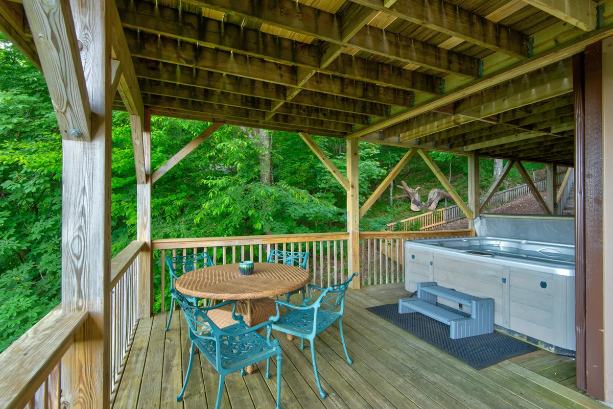 A tranquil wooden deck is shown with a round table for outdoor dining and a hot tub nestled amidst green foliage. The setting suggests intimate moments of relaxation, from stargazing in the hot tub to enjoying morning coffee in the fresh mountain air.