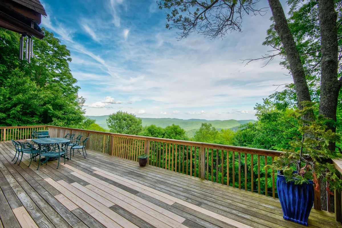 A spacious wooden deck set against a backdrop of lush greenery and distant mountain views. The metal outdoor dining set is arranged for relaxed al fresco meals, while a blue flower pot adds a touch of color and charm. The open, peaceful atmosphere suggests a perfect spot for enjoying sunrise breakfasts or sunset cocktails in nature.