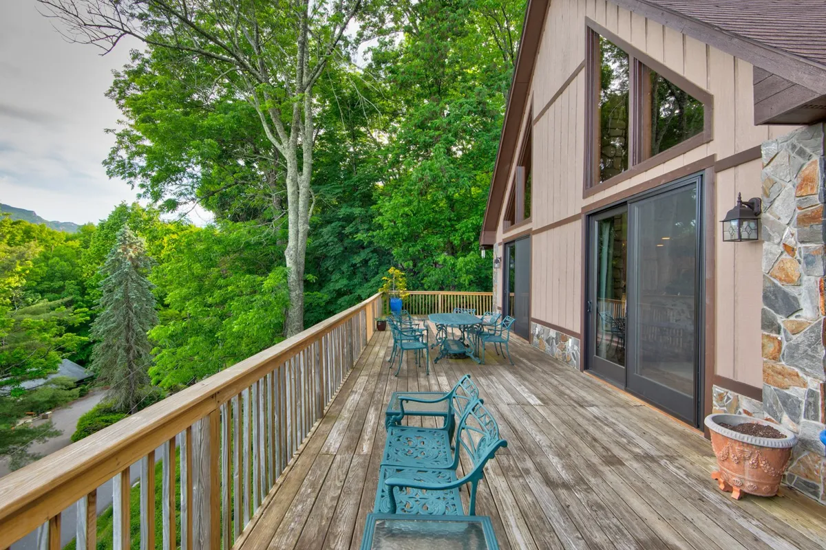 A sweeping view of the home's wraparound deck with inviting seating areas framed by lush trees, all set against a backdrop of majestic mountain vistas. The tranquil scene hints at the deck's optimal positioning for catching the sunrise, sunset, or stargazing, and there is ample space for relaxing or entertaining outdoors.