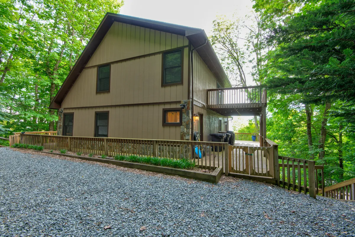 A wide wooden deck stretches out from the house, surrounded by tall trees and natural greenery. Comfortable outdoor furniture provides plenty of space to sit and soak in the views, with a sturdy railing ensuring safety. The deck overlooks a gravel driveway and is enveloped in a serene forest setting, perfectly blending the home with the outdoors.