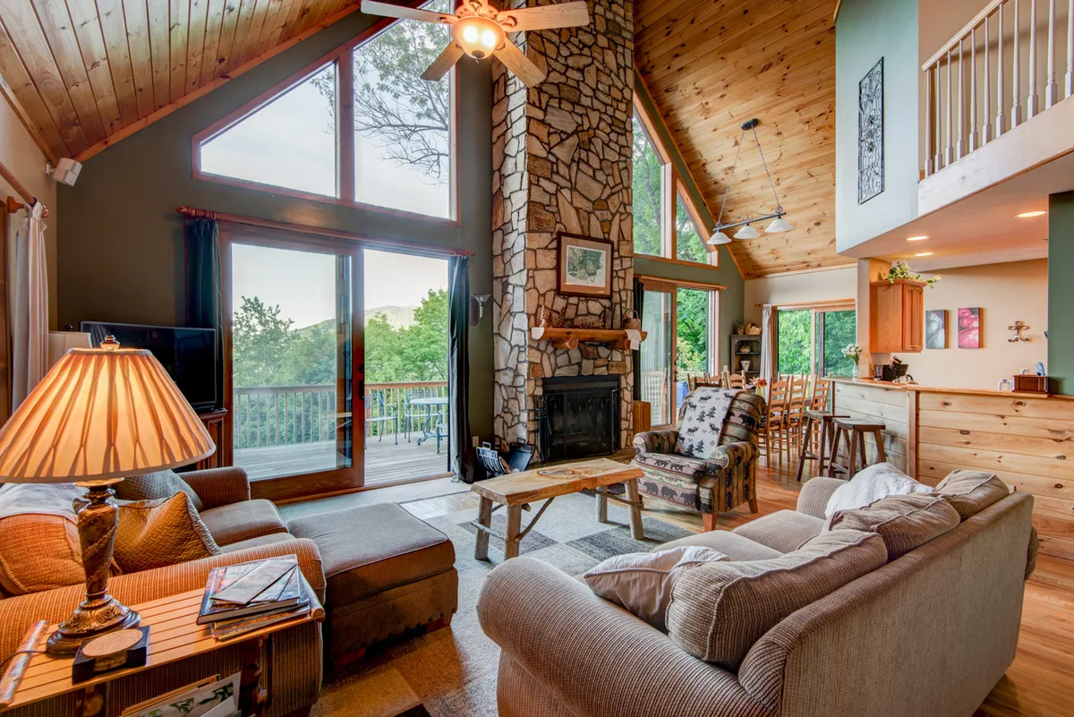 A soaring great room with a dramatic stone fireplace takes center stage, framed by 20-foot wooden-beam ceilings and expansive floor-to-ceiling windows that flood the space with mountain light. Plush, comfortable seating is arranged for easy conversation or relaxing by the fire, making the living area feel both grand and welcoming.