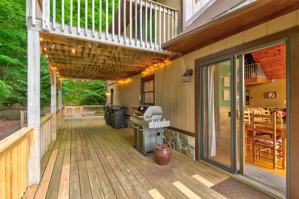 A welcoming exterior deck scene featuring a grill, outdoor dining table, and ample seating, set against a backdrop of lush greenery. The sliding glass door connects the deck directly to the indoors, suggesting seamless transitions between inside and outside activities. The deck is accented with string lights for evening gatherings.
