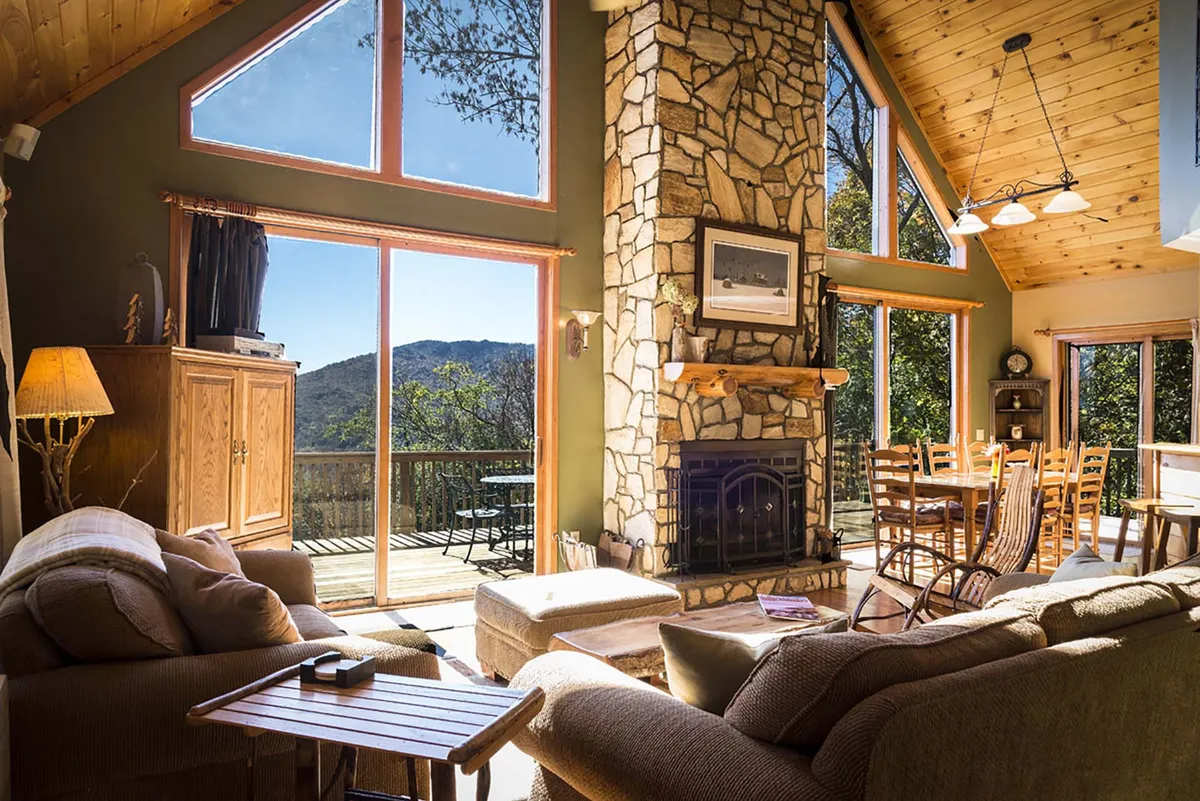 A warm dining area sits adjacent to an imposing stone fireplace, with a rustic wooden table positioned beside expansive windows that open up to views of the mountains. The open-plan layout provides seamless flow for socializing, dining, and relaxing, with natural light enhancing every corner.