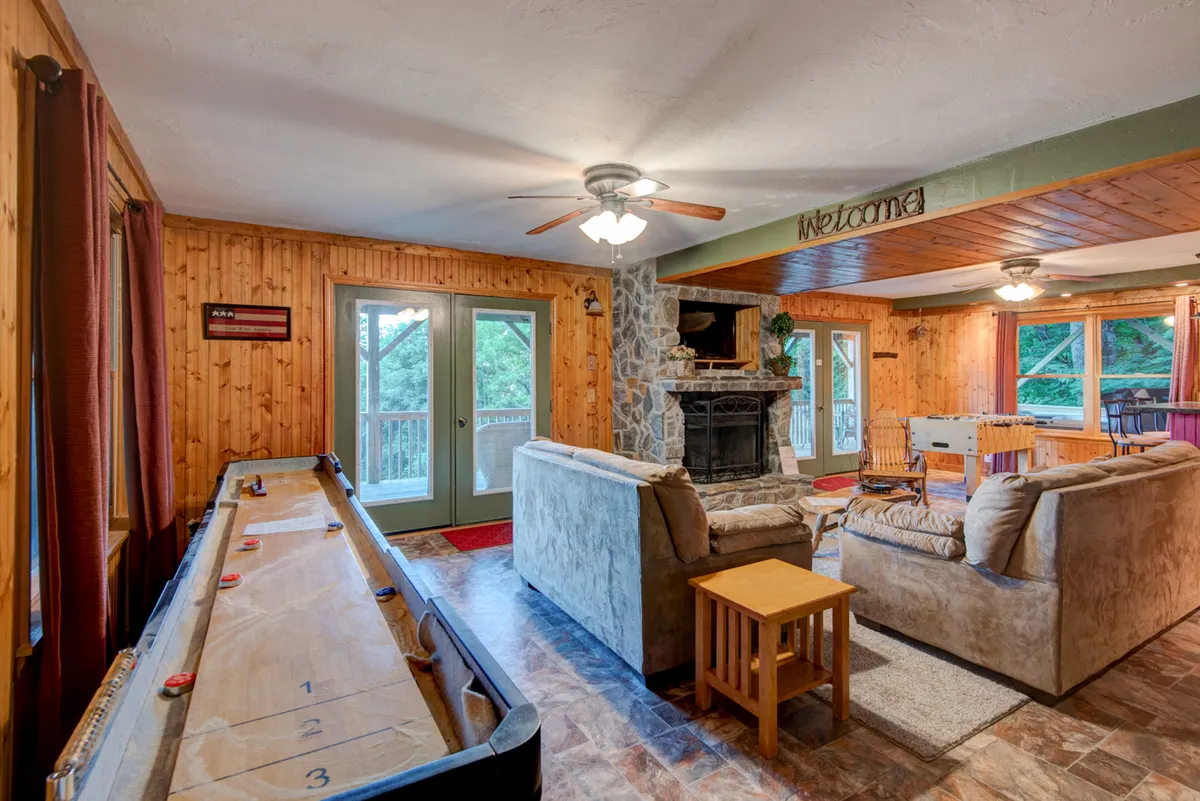This warm, welcoming interior features a comfortable seating area, a fireplace, sliding glass doors, and a shuffleboard table. The multi-functional layout clearly supports relaxing, playing games, and enjoying time together, with both leisure and entertainment options visible in the scene.