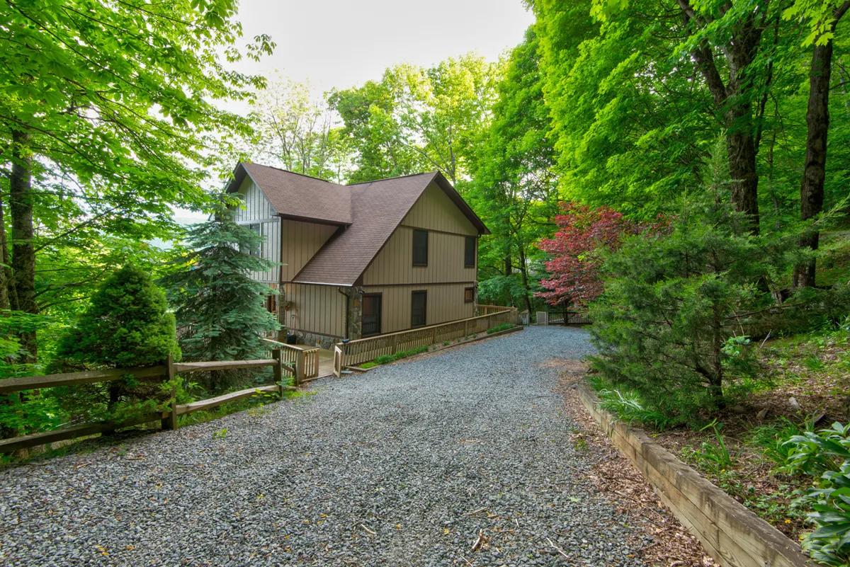 The exterior of the home is shown nestled within vibrant trees and flowering plants. A gravel driveway leads up to the inviting porch, which is easily accessible from the ground level, with no stairs in sight. The scene radiates peace and privacy, with ample space around the home for outdoor walks and adventures.