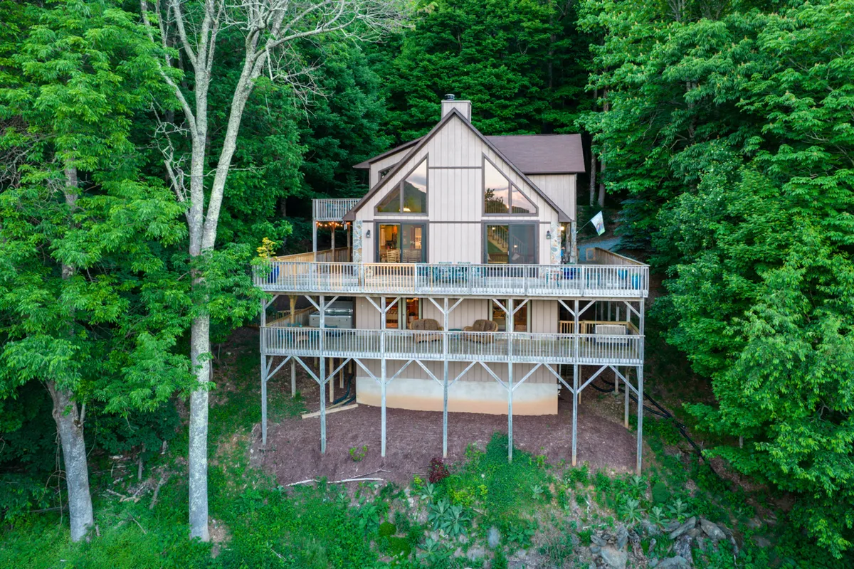 The hero image features a striking single-family mountain home with distinctive triangular architecture, embraced by lush trees. The expansive wrap-around deck is the focal point, perfectly positioned for guests to soak in sweeping long-range mountain views. The peaceful setting is enhanced by soft natural light, inviting guests to imagine relaxing outdoors, hosting gatherings, or enjoying the serenity of nature.