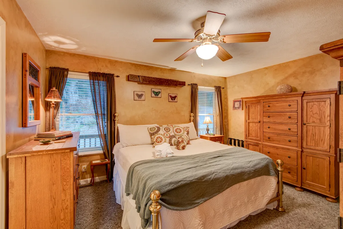 A cozy bedroom with a bed adorned in decorative pillows, a wooden dresser, wardrobe, and cheerful butterfly artwork. Sunlight streams in, creating a warm, inviting atmosphere that suggests restful sleep and peaceful mornings.